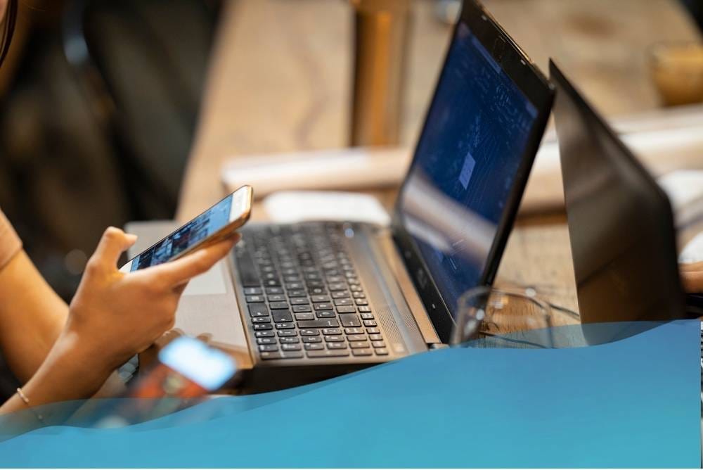 foto di una persona seduta al pc con un telefono in mano e un bicchiere d'acqua sulla scrivania